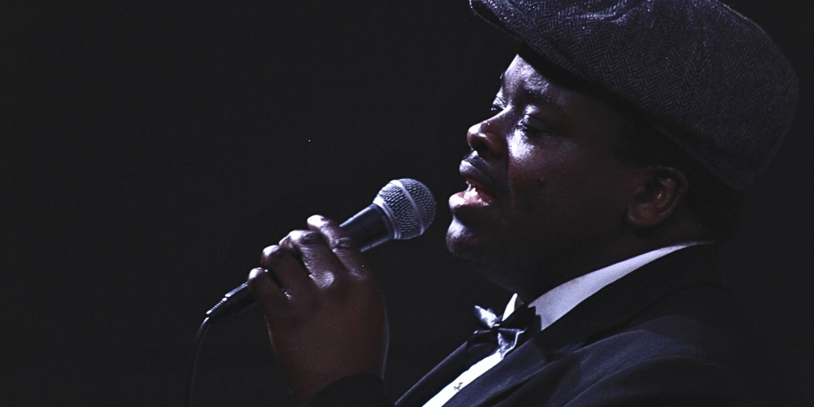 Black man in profile wearing a hat singing into a microphone