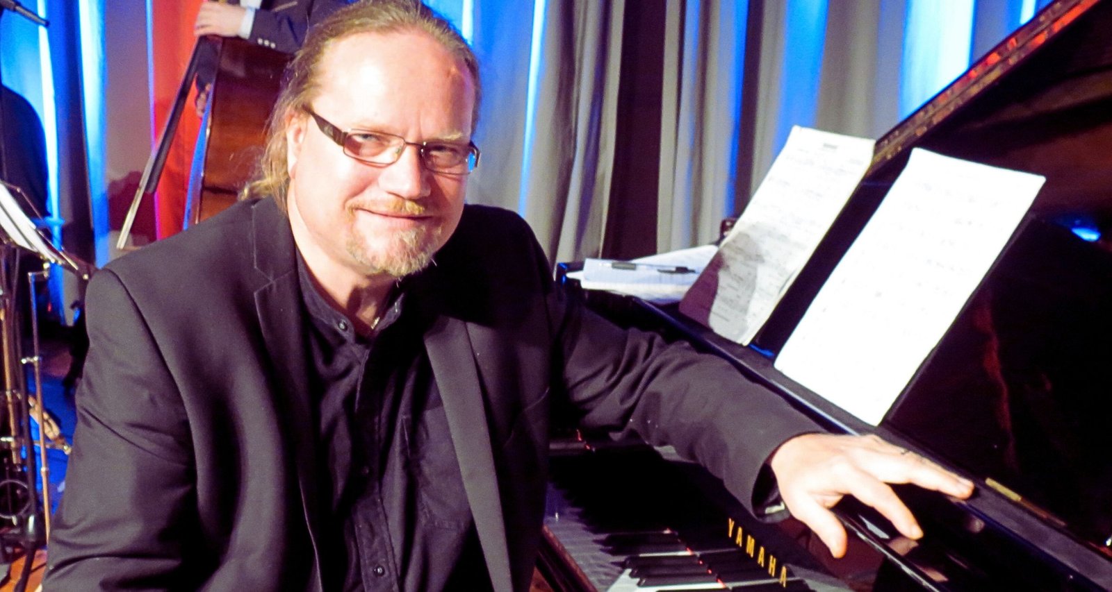 Man wearing glasses in a suit jacket with hands resting on piano facing towardds the camers