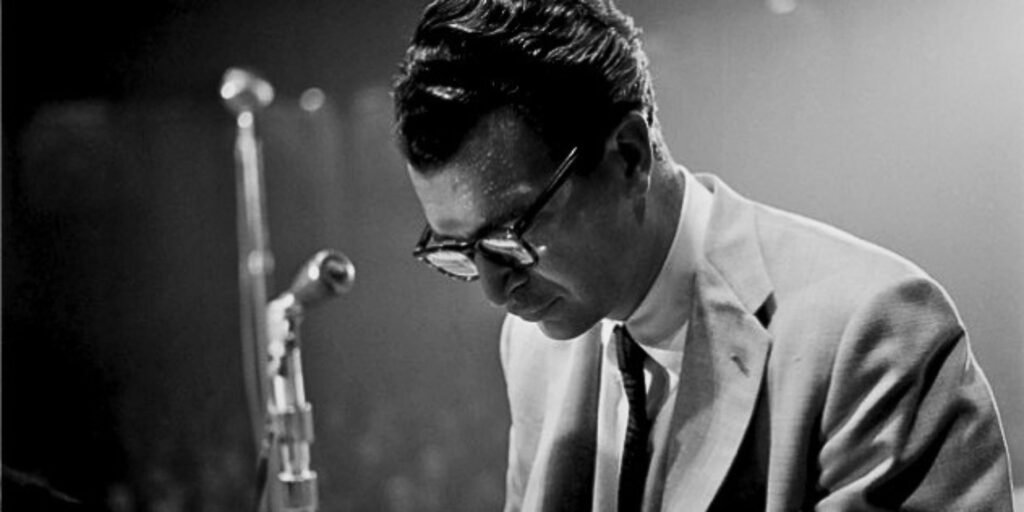 Black-and-white photograph of jazz pianist Dave Brubeck performing on stage, wearing glasses and a light-coloured suit, leaning intently over the piano beneath dramatic concert lighting.