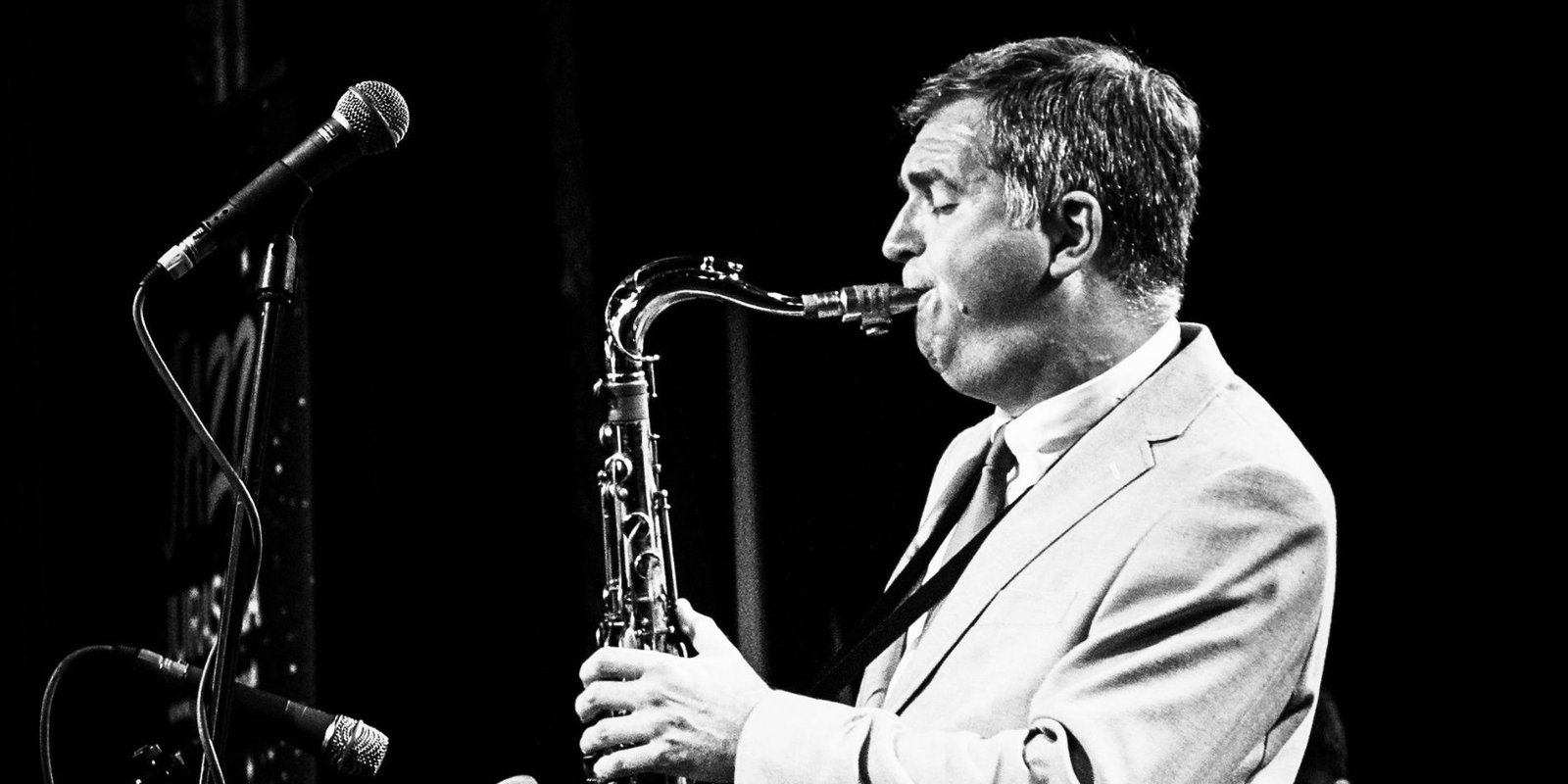 Simon Spillett performing live on tenor saxophone at a jazz concert, standing beside stage microphones in black and white.