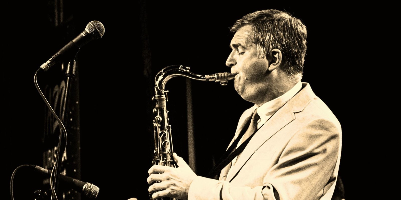 Black and white photo of a jazz saxophonist in a suit playing tenor saxophone on stage beside two microphones.